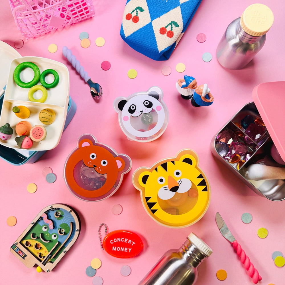 Assorted children's items including a bento box, pacifier, and tins on a pink background with confetti.